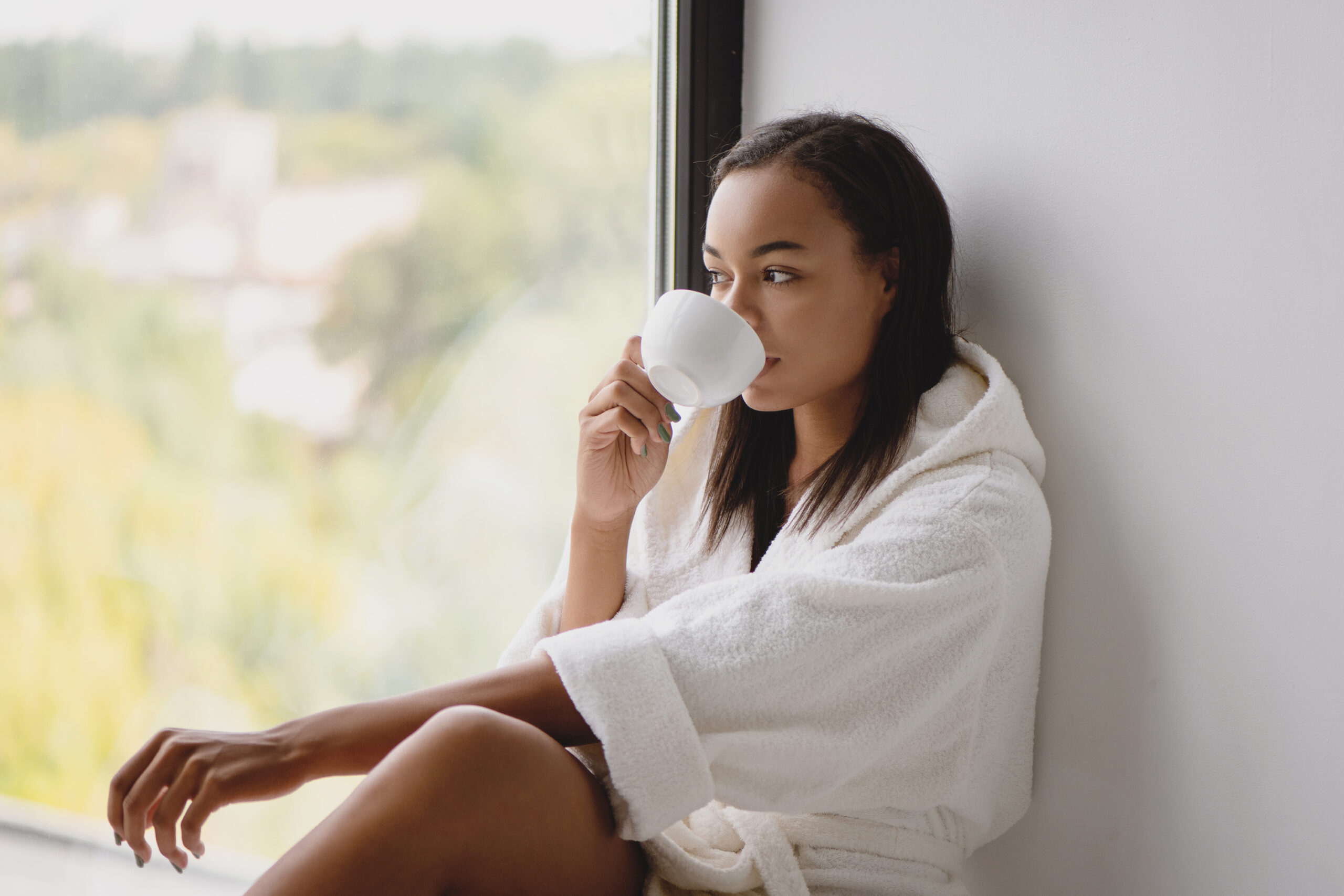 Woman in cozy robe drinking coffee by a window during a calm Sunday reset routine at home.